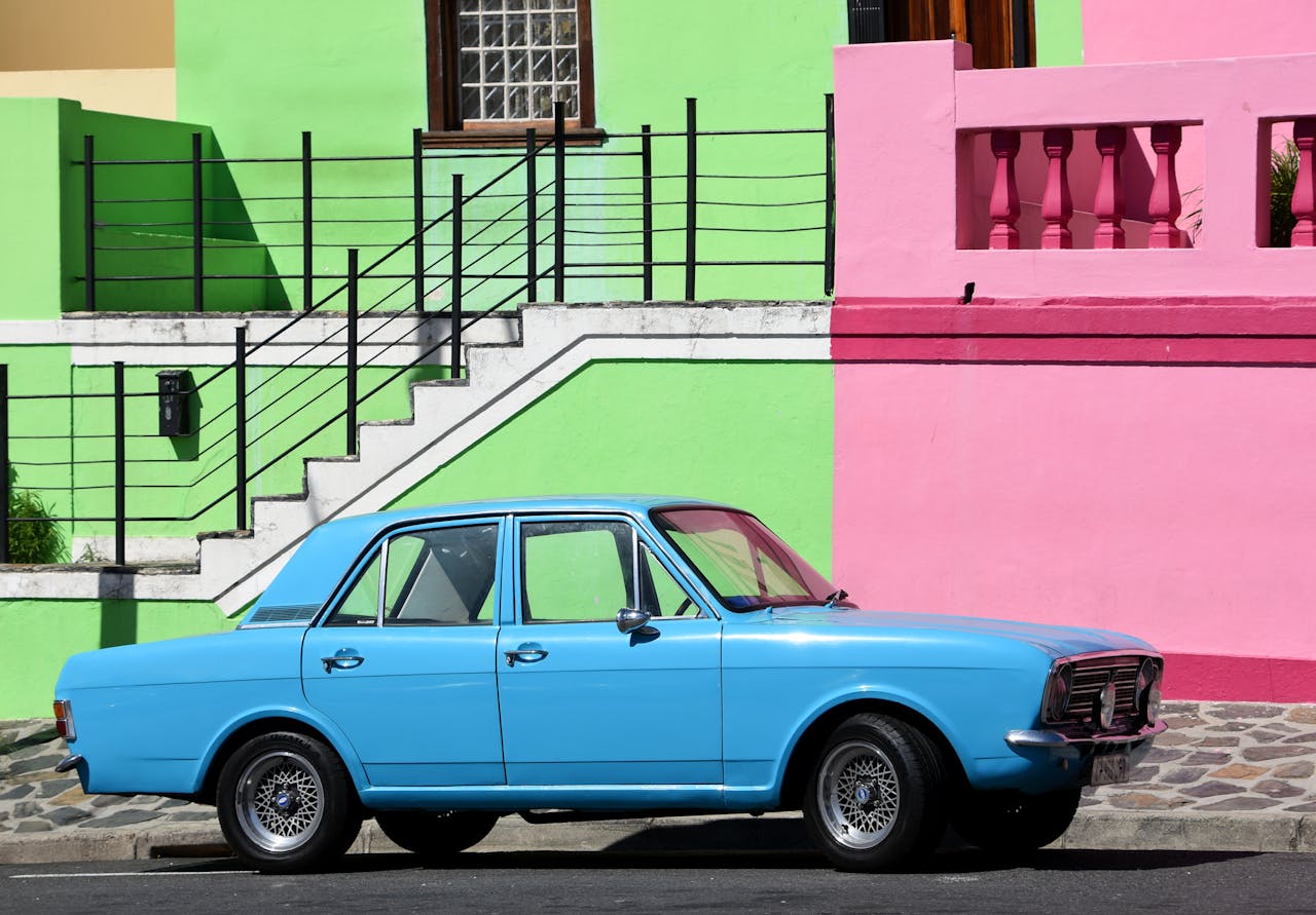 A classic blue car parked beside vibrant green and pink walls in an urban setting.